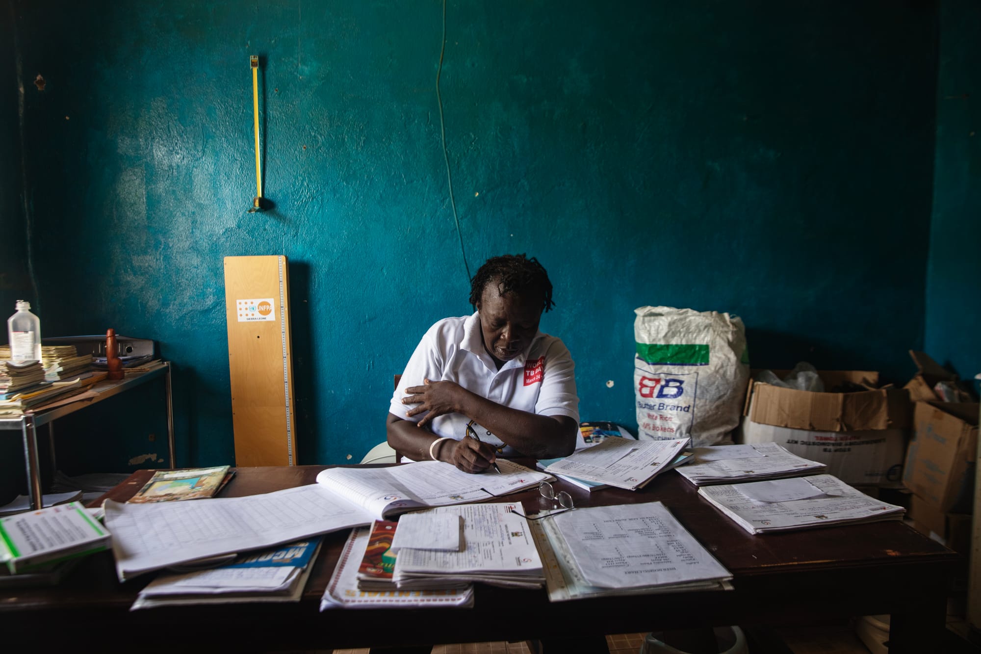 Isatha H. Bangura sits behind a desk, holding a pen and writing on a sheet of paper. 